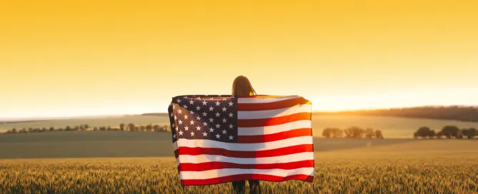 Young woman holding the American flag in a golden wheat field at sunset.
