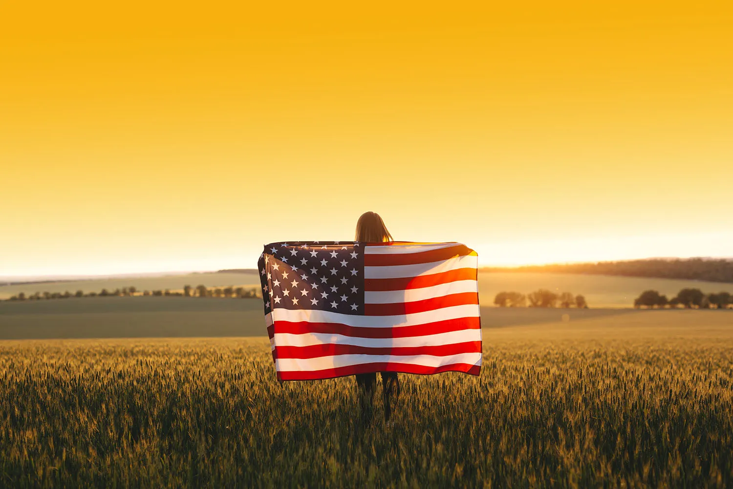 Young woman holding the American flag in a golden wheat field at sunset.