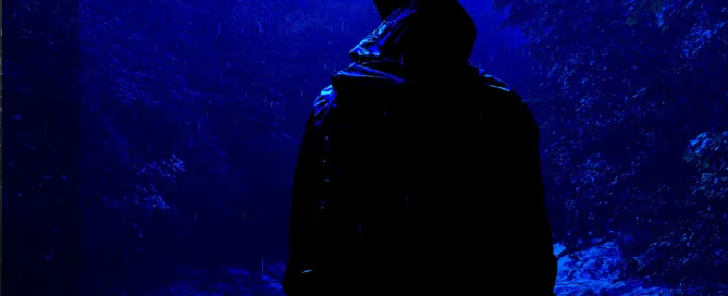 Silhouetted hiker standing alone in a dark, rainy mountain forest at night, overlooking a rushing creek. Atmospheric blue cover for the short story "The Life and Death of an Unimportant Man" by Zac Northup.