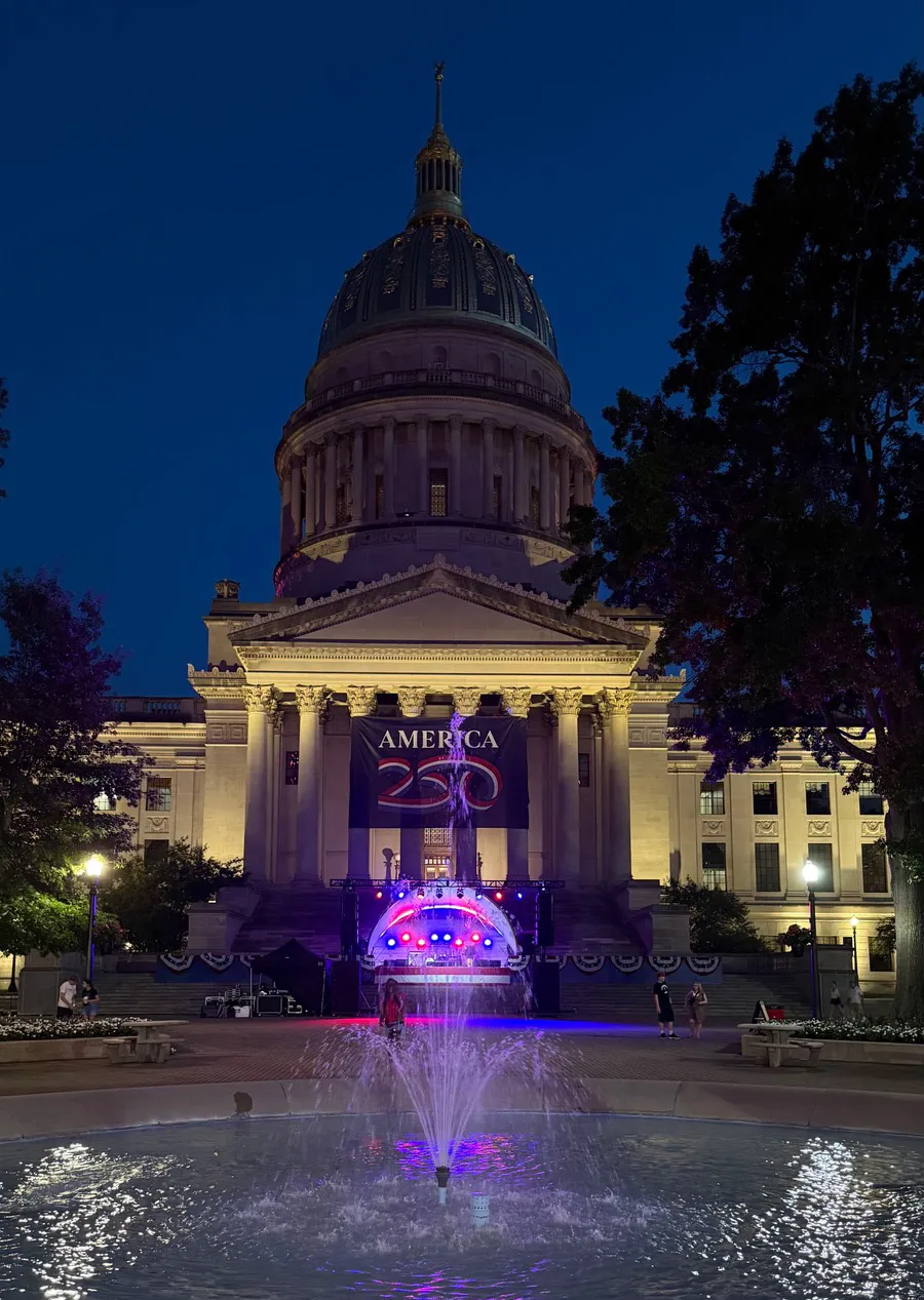 The West Virginia State Capitol at night with an America 250 banner and a lit fountain